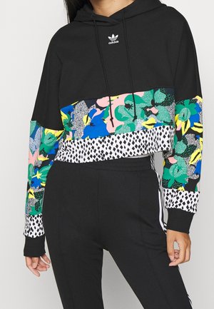 Femme portant un sweat à capuche court Adidas noir avec des motifs floraux et géométriques colorés sur les manches et le torse, assorti à un pantalon noir.