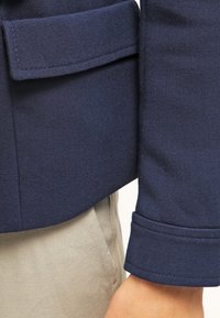 Tmavě modrá bunda z texturované látky s boční kapsou a manžetovými rukávy, doplněná světle zbarvenými kalhotami.