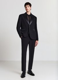 Schwarzer Smoking mit Satinrevers, schmal geschnittene Hose und schwarzes Hemd. Das Material wirkt glatt; polierte schwarze Schuhe komplettieren den Look.