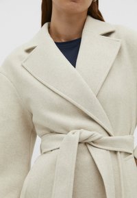 Kobieta ubrana w beżowy wełniany płaszcz z szerokimi klapami i paskiem wiązanym w talii, na ciemnym topie.