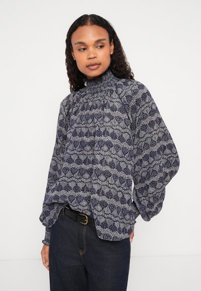 Tengerészkék, hosszú ujjú blúz bonyolult virágmintával; magas redőzött nyakkivágás és dús ujjak. Magas derekú sötét farmernadrággal kombinálva.