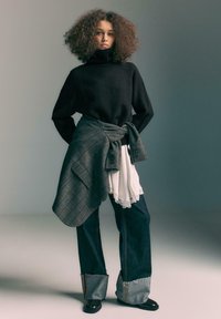 Maglione nero a collo alto, giacca a quadri grigia legata in vita, top bianco stratificato con pizzo, jeans in denim a gamba larga e scarpe nere.