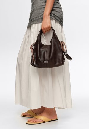 Kobieta trzyma czarną skórzaną torbę na ramię ze sznurkami, ubrana w szary top, długą białą spódnicę i beżowe, płaskie sandały.