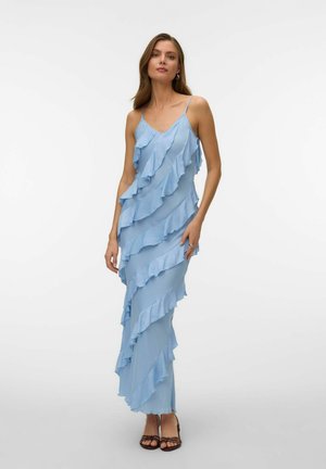 Vrouw die een lichtblauwe mouwloze maxi-jurk draagt met diagonale gerafelde lagen en bruine sandalen met bandjes, staand tegen een witte achtergrond.