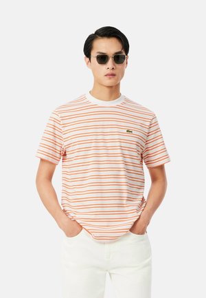 Homme portant des lunettes de soleil, un T-shirt blanc et orange rayé avec un petit logo et un pantalon blanc, debout les mains dans les poches.