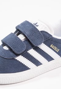 Navy blue suede sneaker with white stripes, Velcro straps, and white rubber sole. "Gazelle" logo in gold on the side.