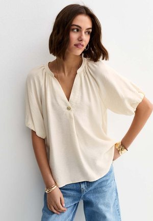 Vrouw met kort bruin haar, gekleed in een losse crèmekleurige blouse met een enkele knoop en lichtblauwe spijkerbroek, aangekleed met gouden armbanden en grote oorringen.