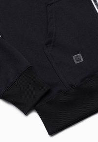 Hoodie noir avec un tissu doux, des poignets côtelés, une poche avant et un petit patch logo près de la zone de la poche.