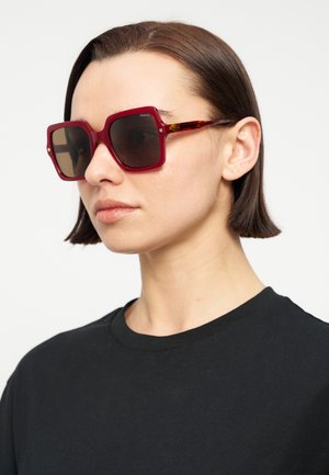 Donna con capelli castani corti che indossa grandi occhiali da sole scuri con montatura rossa e una maglietta nera con collo a giro su sfondo semplice.