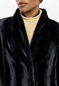 Crni kaput od umjetnog krzna s mekom teksturom, s ovratnikom u obliku šal i dubokim V-izrezom, složen preko bež turtleneck majice.
