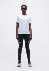 Άτομο που φοράει μαύρο κολάν με αντανακλαστικές ρίγες, ανοιχτό μπλε κοντομάνικο μπλουζάκι, μοβ παπούτσια για τρέξιμο και μαύρα γυαλιά ηλίου, στέκεται μπροστά σε λευκό φόντο.