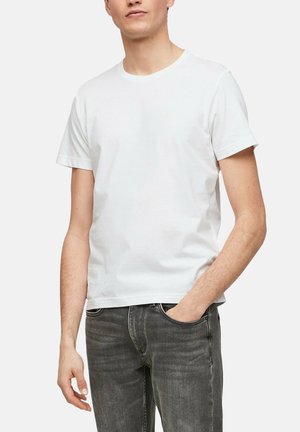 Homme portant un t-shirt blanc uni à manches courtes et un jean noir délavé, avec une main dans la poche sur un fond uni.