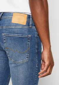 Kék denim farmernadrág, világosbarna címkével a derékpánton, dekoratív öltésekkel díszített hátsó zsebbel, sima textúrával és karcsú szabással.
