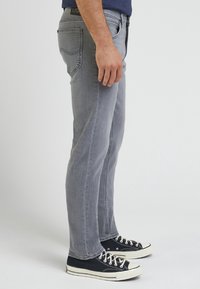 Man wearing gray slim-fit jeans and black low-top canvas sneakers standing sideways on a plain light background.