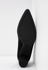 Chaussure à talon haut en daim noir avec un bout pointu, dotée d'une semelle extérieure lisse et d'un léger contour, inclinée sur un fond blanc.