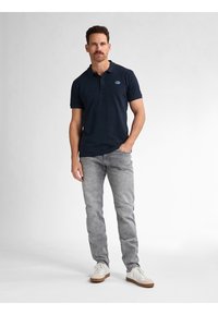 Tummansininen puuvillapolo-paita, jossa on kolme nappia kauluksessa, yhdistettynä vaaleanharmaisiin farkkuihin ja valkoisiin tenniskenkiin. Sujuva pinta, vähäistä brändäystä.