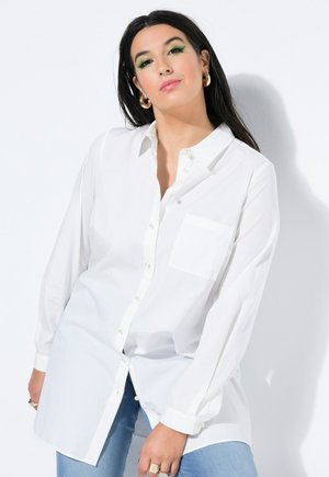 Kvinde med langt mørkt hår, iført grøn øjenskygge, guldfarvede hoops øreringe, en hvid skjorte med knapper og lyseblå jeans mod en hvid baggrund.