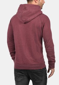 Maroon hoodie made of soft fabric, featuring a large hood, ribbed cuffs, and hem, with a small logo patch near the bottom left.