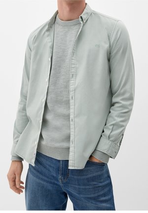 Homme portant une chemise boutonnée gris clair ouverte sur un pull en maille gris et un jean bleu, avec une main dans la poche, sur fond blanc.