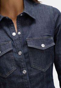 Denimskjorta i mörkblått med knäppning framtill, spetsig krage, två bröstfickor och silverfärgade detaljer. Texturerat tyg upptäckbart.