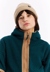 Teal fleecová bunda s hnedým zipsom a kapucňou, s textúrovaným povrchom. Na hlave je béžový rebrovaný čiapka s emblémom.