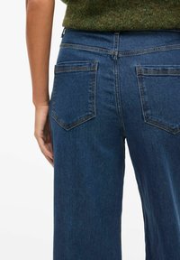 Jeans de mezclilla azul oscuro con un corte relajado, que cuentan con dos bolsillos traseros y costuras visibles. La textura es suave con un ligero brillo.