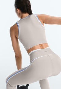 Femme aux cheveux tressés en tenue de sport beige soulevant une kettlebell noire sur un fond clair.