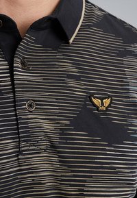 Polo noir avec des rayures horizontales beiges, un plastron à trois boutons, et un écusson logo avec des ailes sur le côté gauche. Texture douce.