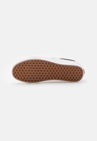Ein einzelner weißer Sneaker liegt auf der Seite und zeigt eine braune Gummisohle mit Waffelmuster vor einem weißen Hintergrund.