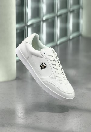 Baskets basses blanches à lacets, avec un logo de crâne noir et blanc sur le côté, flottant au-dessus d'un sol en béton poli près d'un mur en blocs de verre.