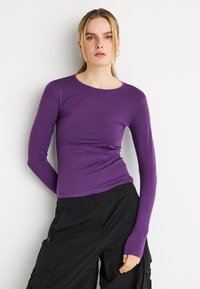 Μωβ εφαρμοστή μπλούζα με μακριά μανίκια κατασκευασμένη από απαλό ύφασμα με στρογγυλή λαιμόκοψη. Διαθέτει ελαφρώς συγκεντρωμένη λεπτομέρεια στη μέση. Φοριέται με μαύρο παντελόνι καρότσα.