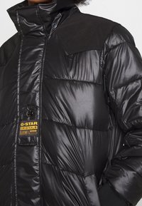Chaqueta acolchada negra con acabado brillante, que presenta un cuello alto, cierre de botones y una etiqueta de tela que dice "G-STAR ORIGINALS RAW DENIM".