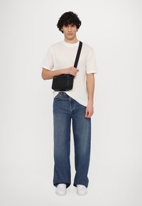 Černá prošívaná crossbody taška se zipovým uzavřením a nastavitelným popruhem. Model má na sobě bílé tričko a široké džíny.