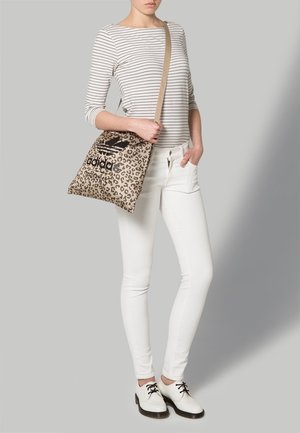 Donna che indossa un top a righe e pantaloni bianchi, mentre tiene una borsa a tracolla Adidas con stampa leopardata, logo nero e tracolla beige.