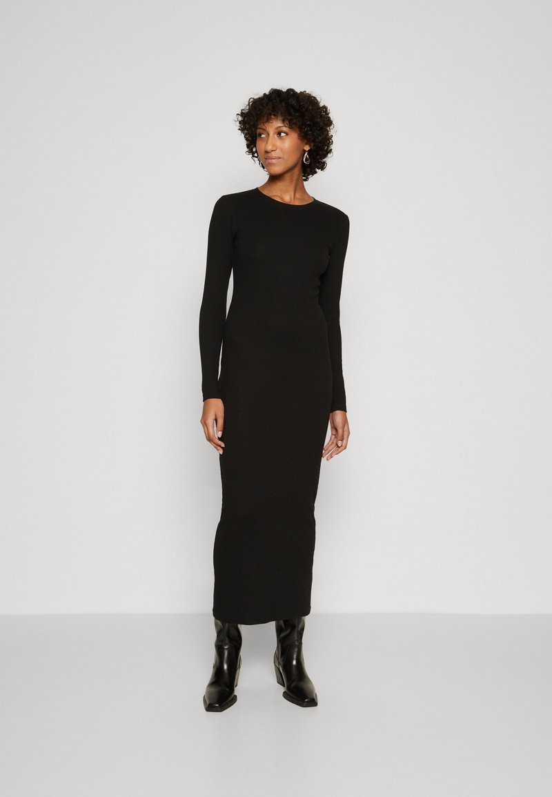 Black long-sleeve dress made of smooth fabric, fitted silhouette, crew neck, and ankle-length design, worn with knee-high black boots.