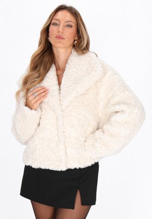 Vrouw met lang golvend haar, gekleed in een witte donzige jas en een zwarte rok met een inkeping, poseert met één hand op de kraag van de jas en de andere op de heup.