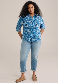 Blå blommig skjorta med knappar och långa ärmar, kombinerad med ljusblå jeans med upprullade benslut och bruna sandaler. Slät tyg med ett livligt mönster.