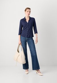 Marineblå knappet skjorte med trekvartlange ermer, kombinert med utsvingte blå jeans, holder en beige hobo-veske og har på seg nude hæler.
