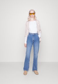 Hochtaillierte hellblaue Jeans mit geradem Bein, kombiniert mit einem weißen Hemd und einem beigefarbenen Leinen-Überhemd. Gelbe Sonnenbrillen akzentuieren den Look.