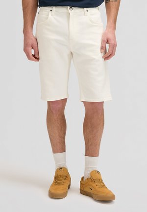 Mężczyzna w kremowych spodenkach do kolan, białych skarpetach oraz beżowych butach Nike, stojący na jednolitym jasnym tle.