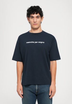 Junger Mann mit lockigem Haar, der ein marineblaues T-Shirt mit dem Text "neanche per sogno" trägt, steht vor einem schlichten weißen Hintergrund.