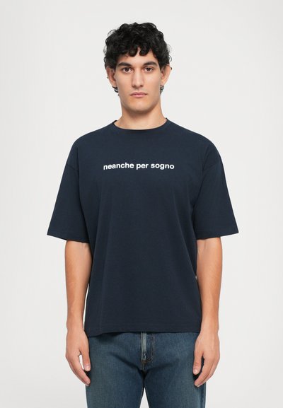 Young man with curly hair wearing a navy blue t-shirt with "neanche per sogno" text, standing against a plain white background.