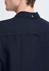 Camicia in lino blu navy con colletto rialzato e un bottone sul retro. La texture appare liscia con una leggera lucentezza.