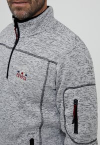 Šviesiai pilkas pliušinis megztinis su tamsiai taškuota tekstūra. Sukurtas su apykakle, užtrauktuku ir logotipu su raudonais, mėlynais ir geltonais akcentais. Šone yra užtrauktuku uždaromas kišenė.