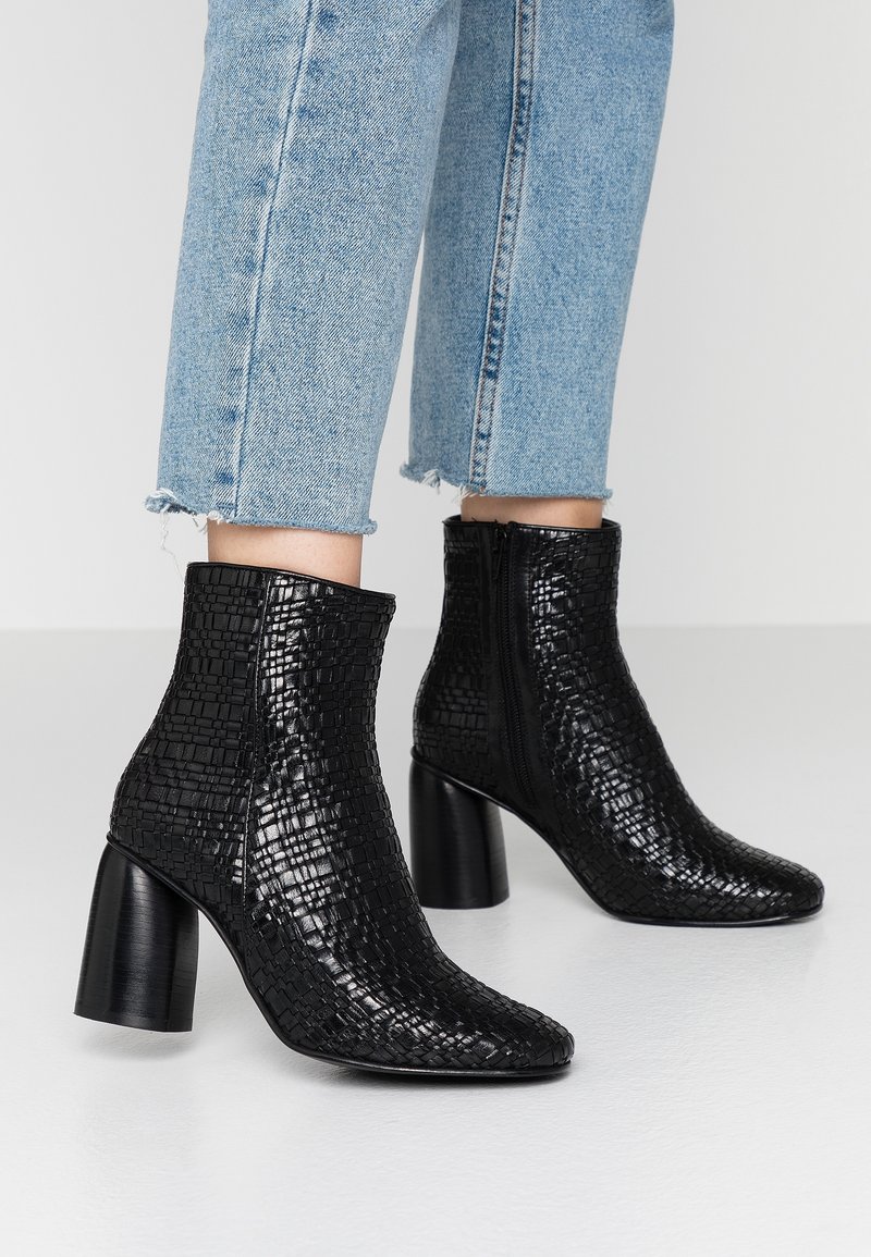 Czarne botki za kostkę z ekologicznej skóry z teksturowanym wzorem krokodyla, spiczaste noski i masywne obcasy, zestawione z przetartymi jasnoniebieskimi dżinsami.