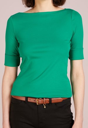 Femme portant une chemise verte ajustée aux manches jusqu'au coude et une ceinture marron sur un pantalon noir, debout devant un fond clair.