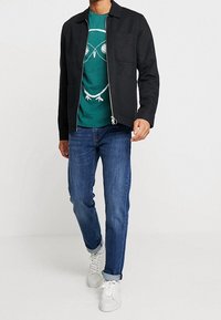 Hombre vestido con una camiseta gráfica de búho en color verde azulado, chaqueta negra con cremallera, jeans azules con puños y zapatillas blancas, caminando contra un fondo liso.