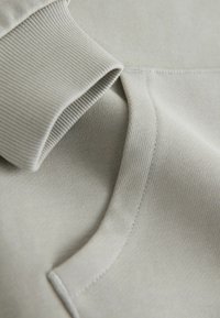 Nahaufnahme eines hellgrauen Stoffs mit geripptem Bündchen und detaillierter Naht auf weichem, strukturiertem Material.