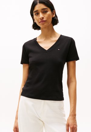 Femme portant un t-shirt noir à col en V et manches courtes et un pantalon blanc, debout devant un fond blanc uni.