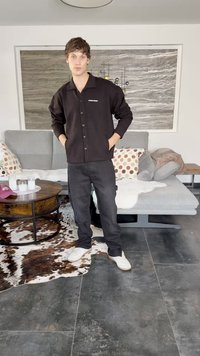 Camicia nera con bottoni e logo, pantaloni cargo neri e sneakers bianche. Sullo sfondo ci sono un divano grigio, un tavolo rotondo in legno e cuscini decorativi.